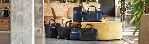 A collection of dark blue and black Floris van Bommel leather travel bags and duffel bags arranged on and around a round yellow ottoman in a modern, industrial-style interior with concrete pillars and wood panelling.