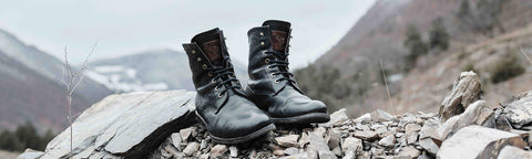 A pair of black floris van Bommel leather lace-up hiking boots resting on a rocky mountain path with a blurred mountain landscape in the background.
