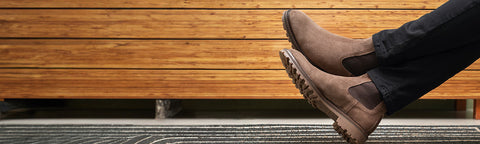 A person's legs wearing brown suede Floris van Bommel Chelsea boots and dark jeans, shown from the side against a wooden wall and textured floor.