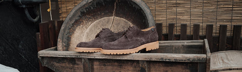 A pair of dark brown suede Floris van Bommel lace-up shoes with tan soles, displayed on a rustic wooden crate in front of a large vintage tire and bamboo wall.