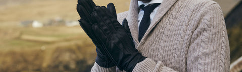 A person wearing a beige cable-knit cardigan, white shirt, black tie, and black Floris van Bommel gloves, standing outdoors with a blurred natural background.