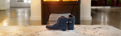 A pair of cozy, dark blue Floris van Bommel knit slipper boots with a soft brown lining, placed on a white textured rug in front of a glowing fireplace.