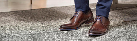 A close-up of a person's feet wearing brown Floris van Bommel leather dress shoes and dark blue trousers, standing on a textured gray carpet.