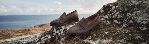 A pair of brown Floris van Bommel suede penny loafers resting on a mossy rock overlooking the ocean, creating a rugged coastal lifestyle scene.