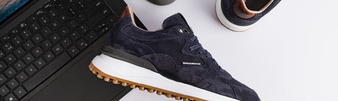 A dark navy blue suede  Floris van Bommel sneaker with a white midsole and a brown outsole, shown in a styled flat-lay composition next to a laptop keyboard.