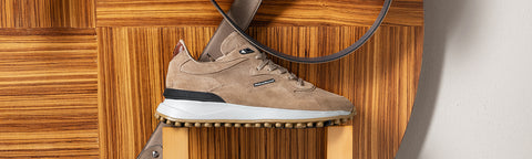 A single low-top Floris van Bommel sneaker in taupe suede, displayed on a wooden riser against a wood-panelled background.