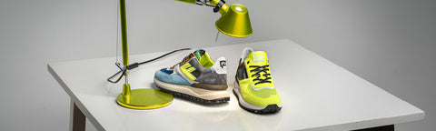 Two athletic Floris van Bommel sneakers displayed on a white table under a green desk lamp against a grey studio background.