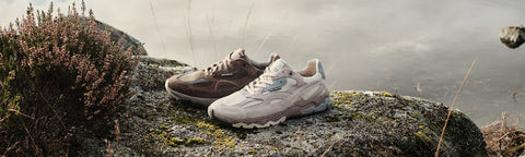 A pair of Floris van Bommel sneakers set on a mossy rock outdoors, with heather plants on the left and a misty natural background behind them.