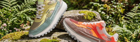 Two athletic Floris van Bommel sneakers are displayed on mossy rocks in a lush outdoor setting with green ferns. One shoe features a light green and beige upper with white soles, while the other features a translucent taupe upper with bright orange and pink accents and white soles.