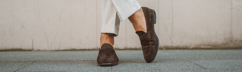 A close-up of a person's legs and feet wearing dark brown suede loafers from Floris van Bommel and off-white trousers, walking on a grey concrete sidewalk against a beige wall.