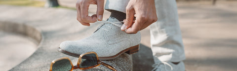 A close-up lifestyle shot of a person tying the laces of a light gray textured suede Floris van Bommel shoe, with their legs in light-colored trousers visible. A pair of sunglasses rests on the concrete surface below.