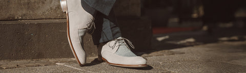 A close-up of a person's legs wearing light grey suede lace-up shoes, brand Floris van Bommel with dark blue jeans, standing on a stone pavement.