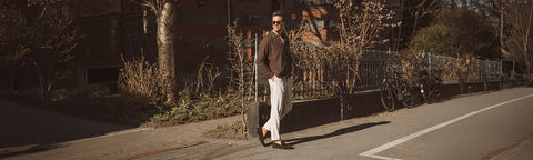 A man walking outdoors on a sunlit street beside a sidewalk, wearing a dark brown jacket, light beige trousers and dark Floris van Bommel shoes.