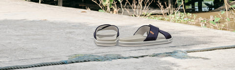 A pair of dark navy slide Floris van Bommel sandals with light gray soles, placed on a concrete surface outdoors.