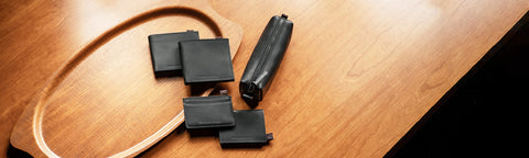 Four black rectangular wallets and a black cylindrical component are arranged on a wooden surface next to a wooden oval tray.