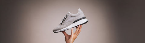 A light grey athletic Floris van Bommel sneaker with black and white details, held up by a person's hand against a dark gray gradient background.