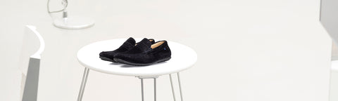 A pair of black Floris van Bommel slip-on loafers displayed on a small round white table in a minimalist white interior setting.