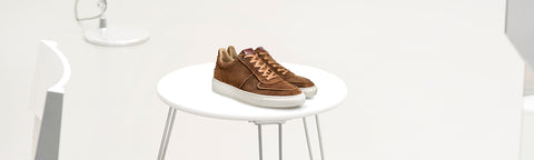 A pair of brown suede low-top van Bommel sneakers with white soles, displayed on a white round table in a minimalist white room.