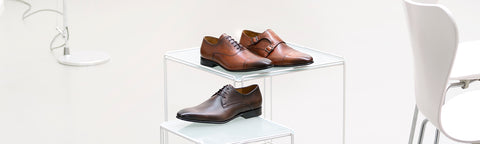 Two pairs of brown leather van Bommel dress shoes are displayed on clear acrylic risers in a bright, minimalist room with a white chair and lamp visible in the background.