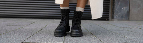 A close-up of a person's legs wearing black lace-up Floris van Bommel boots with white socks, standing on a grey stone pavement in front of a dark metal shutter.