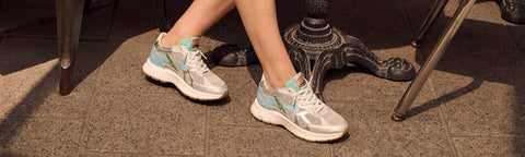 A person wearing light gray and mint green Floris van Bommel sneakers, seated outdoors on a stone floor near a metal table base.