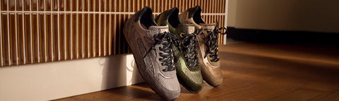Three athletic Floris van Bommel sneakers in grey, olive green, and beige tones leaning against a slatted wall on a wooden floor.