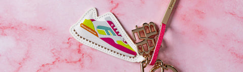 A close-up of a colourful Floris van Bommel keychain featuring a white silhouette-shaped charm with pink details, gold-tone metal keyrings, and a pink strap, set against a pink marble background.