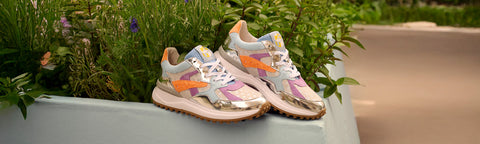 A pair of colourful Floris van Bommel sneakers resting on the edge of a planter box outdoors, with green foliage and a blurred path in the background.