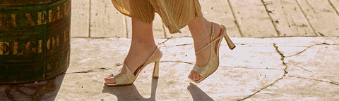 A close-up lifestyle shot of a person wearing beige Floris van Bommel strappy high-heeled sandals with a gold pleated skirt, walking on a sunlit stone pavement next to a barrel.