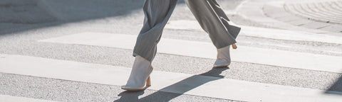 A person walking across a crosswalk outdoors, shown from the knees down, wearing light grey wide-leg trousers and white Floris van Bommel ankle boots with gold heels.