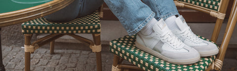 White Floris van Bommel sneakers worn with light blue jeans, shown in a real cafe-like environment with woven chairs and a table visible.