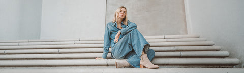A woman sits on concrete steps wearing a long-sleeve denim jacket and matching denim skirt, with beige Floris van Bommel ankle boots, against a minimalist concrete wall.