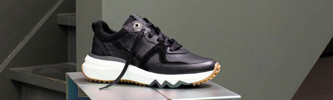 A black Floris van Bommel sneaker with a chunky white midsole and a tan outsole, displayed on a grey platform against a dark gray background.
