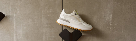 A white Floris van Bommel sneaker with light grey overlays and a gum sole, displayed on a black stand against a textured gray concrete wall.
