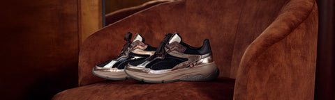 A pair of Floris van Bommel sneakers with metallic silver and black panels, resting on a brown velvet armchair in a dimly lit interior setting.