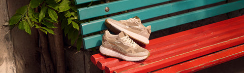 A pair of beige and white Floris van Bommel shoes placed on a red wooden bench next to a turquoise wall, with green leaves hanging on the left.