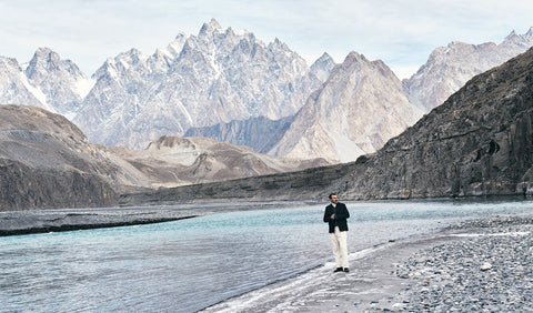 Floris van Bommel stands on a rocky riverbank in a vast mountain landscape, with steep grey and brown peaks rising behind a turquoise river under a pale sky.
