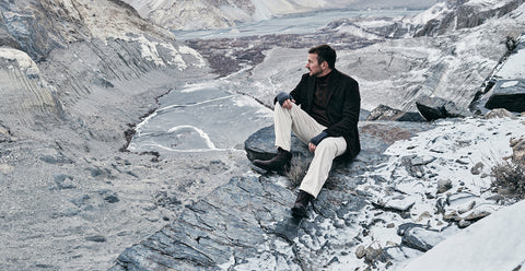 Floris van Bommel sits on a dark stone in a rugged, snow-dusted mountain landscape, wearing a black coat, dark brown turtleneck, cream trousers, and black boots.
