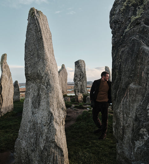 Floris van Bommel in a dark jacket and brown sweater walks between large standing stones in an outdoor stone circle landscape under a pale sky.