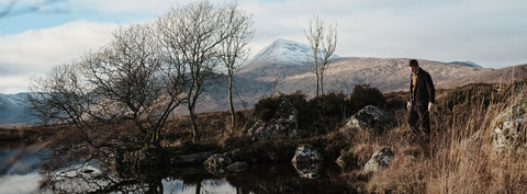 Floris van Bommel stands on a grassy hillside near a reflective pond, with bare trees, rocks, and a snow-capped mountain in the background under a cloudy sky.