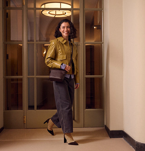 A woman stands in a hallway wearing a shiny mustard yellow leather jacket, dark grey wide-leg pants, black Floris van Bommel heels, and carrying a brown crossbody bag.