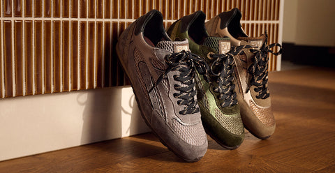 Three athletic sneakers leaning against a slatted wooden wall on a wood floor. The shoes are shown in three colorways: gray, olive green, and beige/tan, all with black laces. Floris van Bommel