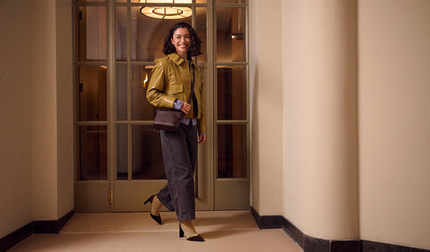 A woman walking through a hallway wearing a shiny olive green jacket, dark grey trousers, and black Floris van Bommel heels, carrying a brown shoulder bag.