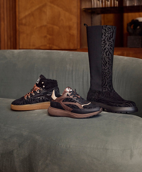 Three fashion footwear pieces from the brand Floris van Bommel displayed on a grey upholstered bench in an indoor setting: a black high-top sneaker with tan and brown laces, a black low-top sneaker with brown and beige accents, and a tall black patterned boot.