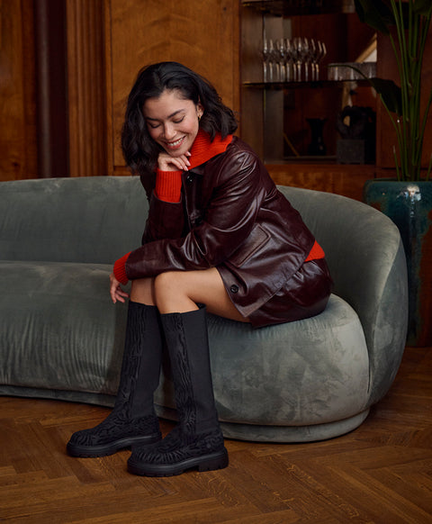 A woman sits on a curved gray sofa in a warm wood-paneled room, wearing a dark brown leather jacket over a bright red sweater, black knee-high lace-up Floris van Bommel boots.