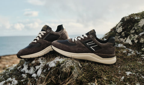 A pair of dark brown Floris van Bommel suede and leather sneakers with white laces and off-white soles, styled on a mossy rock outdoors with the sea and cloudy sky in the background.
