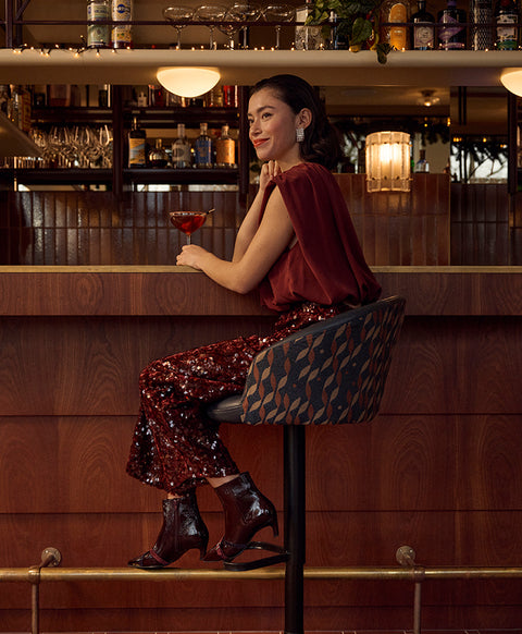 A woman sits on a patterned bar stool at a wooden bar, wearing a burgundy velvet top and matching sequined pants with dark Floris van Bommel ankle boots, holding a glass.