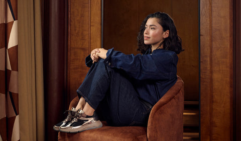 A woman sits in a brown upholstered armchair in a wood-panelled room, wearing a dark navy blue sweatshirt and jeans with black and Floris van Bommel sneakers.
