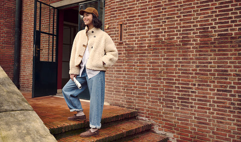 A woman wearing a beige sherpa jacket, white t-shirt, light blue jeans, Floris van Bommel shoes and a brown cap stands on brick steps in front of a red brick building.
