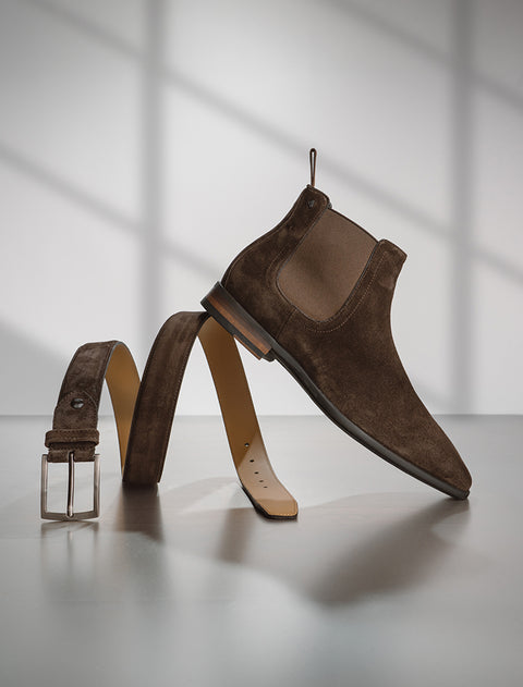 A brown suede Floris van Bommel Chelsea boot displayed in a creative, sculptural pose with its matching belt arranged in a loop behind it, set against a minimalist grey background with soft window shadow patterns.